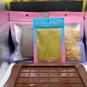 May include: A brown silicone chocolate mold with 16 squares, a pink resealable bag with a chocolate bar inside, a blue resealable bag with a green and yellow bar inside, and a pink resealable bag with a pile of thin, light brown strands inside.