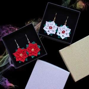 May include: Crochet poinsettia earrings in black boxes. The earrings have red and white flower designs with green leaves and gold-coloured accents. The earrings are displayed in black gift boxes, against a black background.