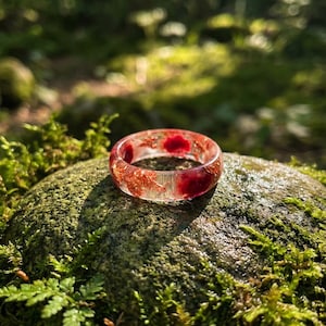 May include: A clear resin ring with red and gold inclusions, resting on a moss-covered rock. The ring's design features a band of clear resin with embedded red and gold elements. The background is a blurred forest scene with sunlight.
