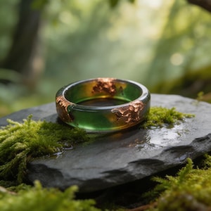 May include: A handcrafted ring with a transparent green resin band, embedded with copper flakes. The ring sits on a dark gray stone, surrounded by green moss, with a blurred forest background. The ring's design evokes a natural, organic aesthetic.
