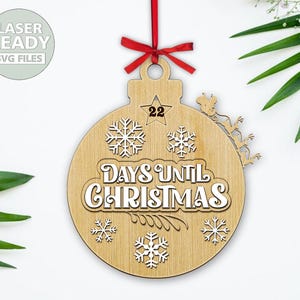 Può includere: Un ornamento natalizio in legno a forma di sfera con il testo "Days Until Christmas" e il numero "22" al centro. L'ornamento è decorato con fiocchi di neve e una stella.