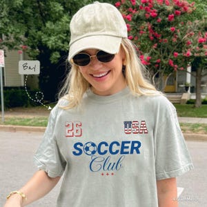 May include: A light green short-sleeve t-shirt featuring the text 26, USA with a flag pattern, and SOCCER Club with a soccer ball graphic. The shirt is paired with a light green baseball cap and round sunglasses.