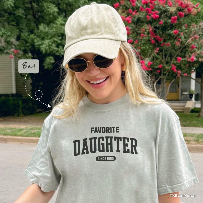 May include: A light green t-shirt with the words "FAVORITE DAUGHTER" printed in black, with "SINCE 1985" underneath. The person is wearing a light green cap and sunglasses. The image is taken outdoors.