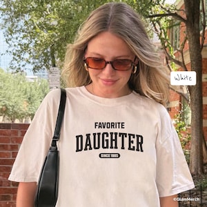May include: A white t-shirt with the words "FAVORITE DAUGHTER" in bold black letters, with "SINCE 1985" underneath. The shirt is worn by a person wearing brown-tinted glasses and gold hoop earrings. A black purse hangs from the shoulder.
