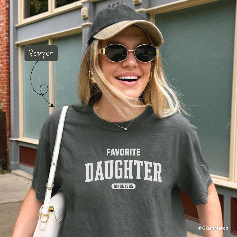 May include: Dark gray t-shirt with the text "FAVORITE DAUGHTER SINCE 1985" in white. The person in the image is wearing a baseball cap, sunglasses, and a white purse. The background includes a building with windows.