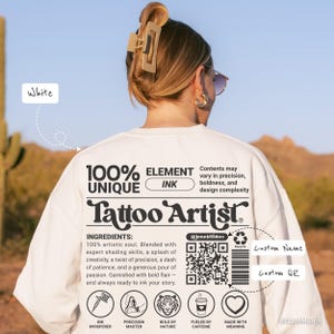 Könnte beinhalten: Ein weißes Sweatshirt mit dem Text "Tattoo Artist" und den unten aufgeführten Zutaten. Das Design umfasst einen QR-Code, Symbole und die Worte "100% Unique Ink". Das Model trägt eine Haarspange und eine Sonnenbrille.