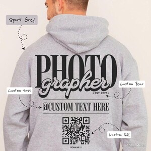 Puede incluir: Sudadera gris deportiva con la palabra "PHOTOGRAPHER" en letras negras grandes en la espalda. Incluye el texto "@CUSTOM TEXT HERE" y un código QR. La sudadera también tiene el texto "+ EST. 2026 +" y las palabras "Custom Year" y "Custom QR".