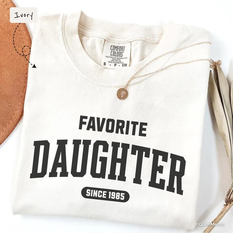 May include: Ivory-colored t-shirt with the words "FAVORITE DAUGHTER" in bold black letters, with "SINCE 1985" below. A gold necklace with a pendant rests on the shirt. The shirt is folded and displayed on a white surface.