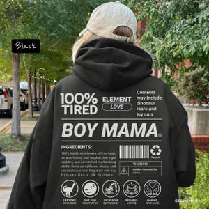 Pode incluir: Moletom preto com a frase branca "100% TIRED BOY MAMA". A parte de trás do moletom inclui uma lista de "ingredientes" e ícones. A pessoa que usa o moletom também está usando um boné de beisebol bege.