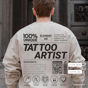 May include: A beige sweatshirt with the words "TATTOO ARTIST" in large black letters. The design includes text that reads "100% UNIQUE" and "ELEMENT INK". Below the text are illustrated icons and the words "INK WHISPERER", "PRECISION MASTER", "BOLD BY NATURE", "FUELED BY CAFFEINE", and "MADE WITH MEANING".