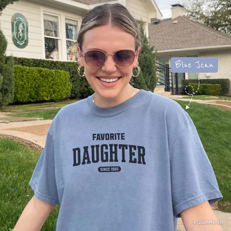 May include: A blue t-shirt with the words "FAVORITE DAUGHTER SINCE 1985" printed in black. The shirt is a light blue color and is being worn outdoors. The person is wearing sunglasses and hoop earrings.