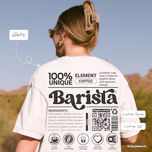 Op de afbeelding: Wit T-shirt met een barista-ontwerp. Het shirt toont de tekst "100% UNIQUE COFFEE Barista" en koffie-gerelateerde pictogrammen. De achterkant van het shirt heeft een ingrediëntenlijst en een QR-code. Het shirt wordt gedragen door een persoon met een zonnebril en een haarclip.
