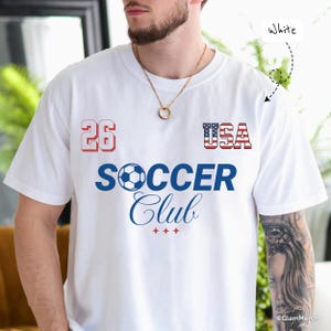 May include: A white short-sleeve t-shirt featuring the number 26 in red, the text USA with a flag pattern, and the blue text SOCCER Club with a soccer ball icon replacing the letter O. Three small red stars are printed below the text.