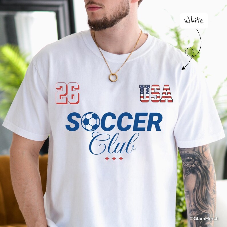 May include: A white short-sleeve t-shirt featuring the number 26 in red, the text USA with a flag pattern, and the blue text SOCCER Club with a soccer ball icon replacing the letter O. Three small red stars are printed below the text.