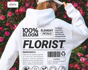 Peut inclure: Un sweat à capuche blanc avec le mot "FLORISTE" en grandes lettres noires. Le dos du sweat à capuche présente un texte sur les ingrédients d'un fleuriste, y compris l'amour des fleurs et la caféine. Le sweat à capuche est entouré de fleurs roses.