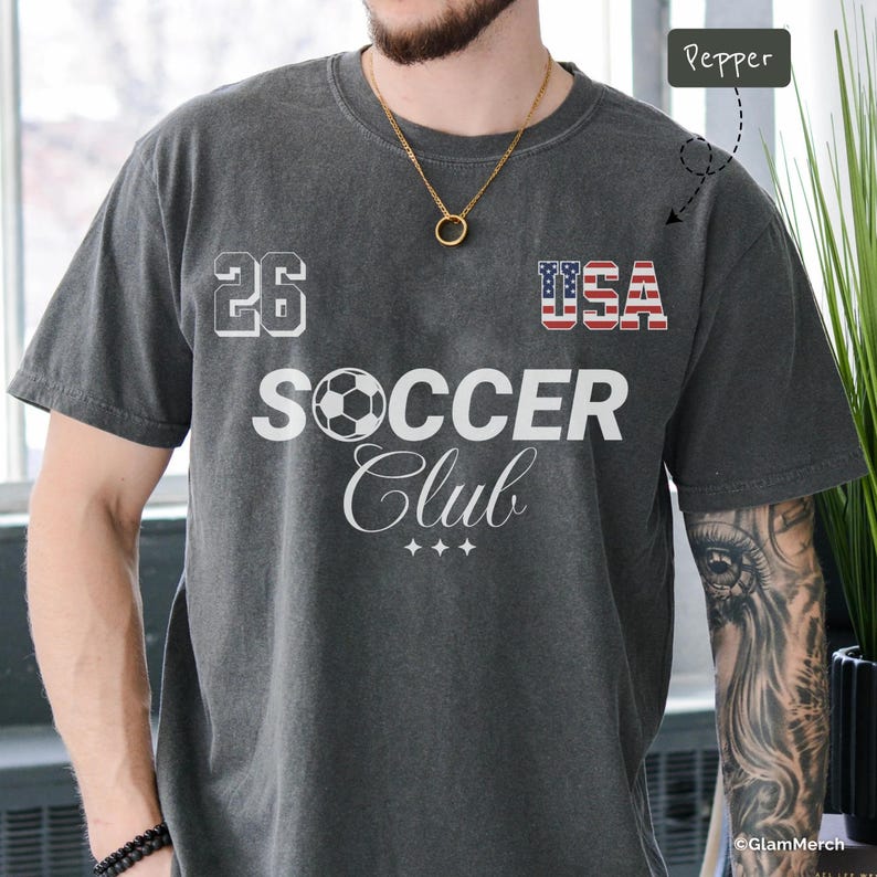 May include: A charcoal gray t-shirt featuring the text "26" and "USA" with an American flag pattern. Below, the word "SOCCER" is printed with a soccer ball replacing the letter "O," followed by the word "Club" in script and three small stars.