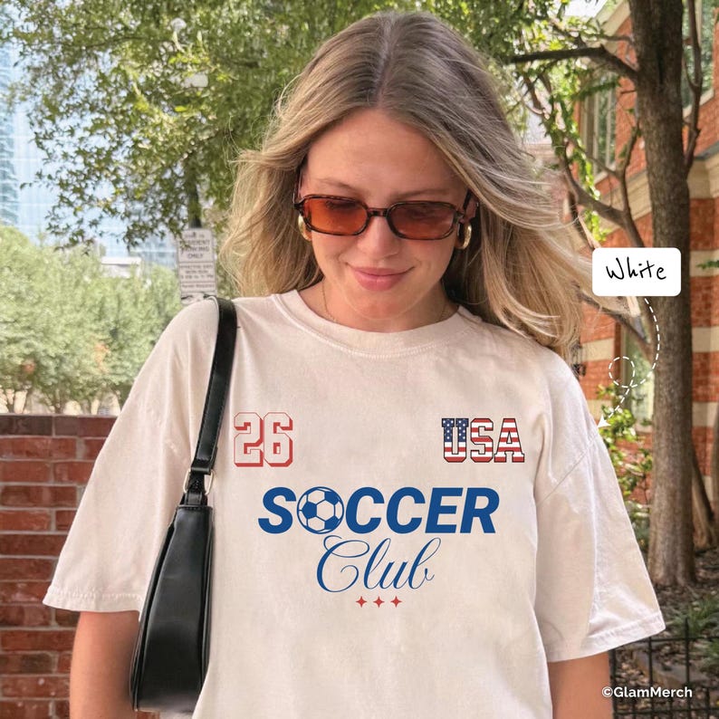 May include: A white short-sleeve t-shirt featuring the text 26 in red, USA in a red, white, and blue flag pattern, and the words SOCCER Club in blue. A soccer ball graphic replaces the letter O in SOCCER, with three small red stars below the text.