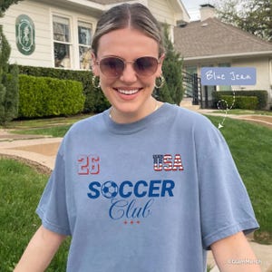 May include: A blue jean colored t-shirt featuring the text 26, USA with a flag pattern, and SOCCER Club with a soccer ball graphic. The shirt has a crew neckline and short sleeves, with a casual fit and a soft, washed-out appearance.