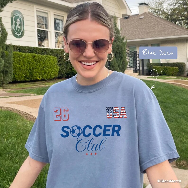 May include: A blue jean colored t-shirt featuring the text 26, USA with a flag pattern, and SOCCER Club with a soccer ball graphic. The shirt has a crew neckline and short sleeves, with a casual fit and a soft, washed-out appearance.
