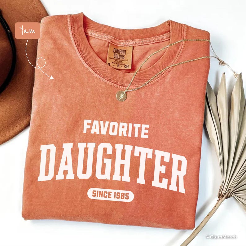 May include: A coral-colored t-shirt with the words "FAVORITE DAUGHTER" printed in white, along with "SINCE 1985". A gold necklace with a pendant rests on the shirt. The shirt is from Comfort Colors.