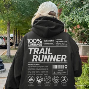 May include: Black hoodie with the text "TRAIL RUNNER" in white. The back of the hoodie features a list of ingredients and icons. The hoodie also has the text "100% COURAGE" and "ELEMENT ENDURANCE". A beige cap is worn.