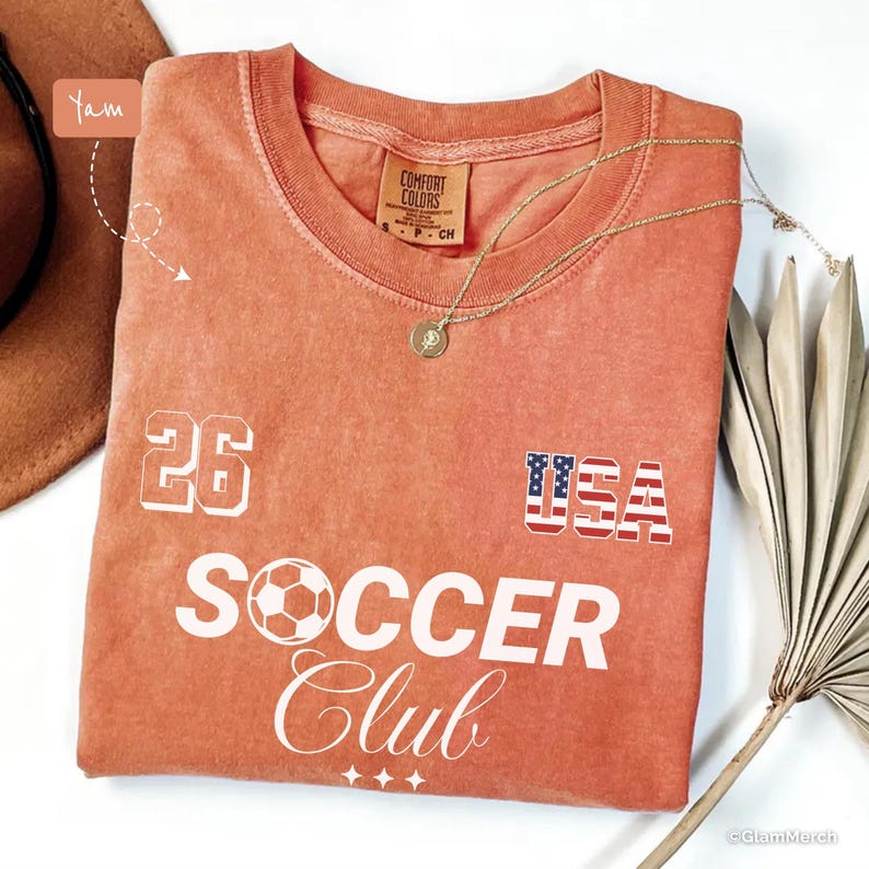 May include: A terracotta-colored short-sleeve t-shirt featuring the white text "26" and "SOCCER CLUB" with a soccer ball graphic replacing the letter "O". The design includes a small "USA" graphic with a flag pattern and three stars.