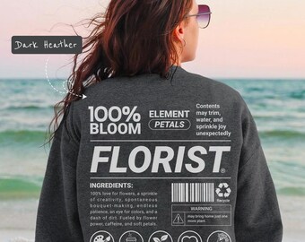 Op de afbeelding: Donkergrijs sweatshirt met het woord "FLORIST" in witte letters. Het sweatshirt heeft een lijst met "INGREDIËNTEN" en pictogrammen voor kleurexpert, cafeïne, plantenverzamelaar, gemaakt met liefde en boeketkunstenaar.