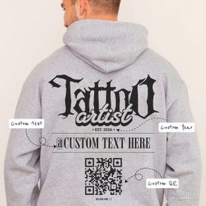 May include: A light grey hooded sweatshirt featuring black text on the back that reads Tattoo artist, EST. 2026, and @CUSTOM TEXT HERE. Below the text is a black QR code with the words SCAN ME underneath. The design includes labels for custom text.