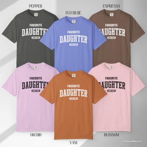 May include: A collection of six short-sleeved t-shirts in various colors, including gray, blue, brown, pink, and orange. Each shirt features the text "FAVORITE DAUGHTER" in a bold, white, collegiate-style font, with "SINCE 1985" below.