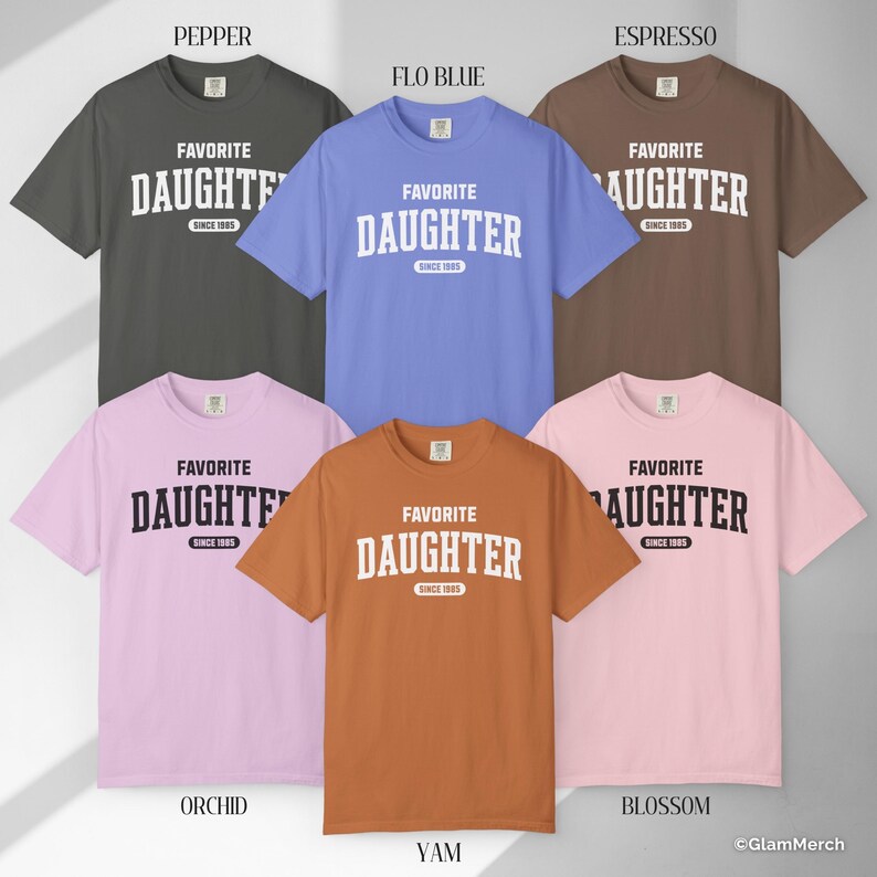 May include: A collection of six short-sleeved t-shirts in various colors, including gray, blue, brown, pink, and orange. Each shirt features the text "FAVORITE DAUGHTER" in a bold, white, collegiate-style font, with "SINCE 1985" below.