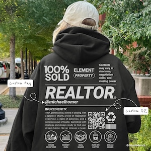 Op de afbeelding: Zwarte hoodie met het woord "REALTOR" in het wit, samen met de tekst "100% SOLD" en "ELEMENT PROPERTY". De achterkant van de hoodie bevat ook de tekst "@michaelhomer" en een lijst met ingrediënten. Een aangepaste QR-code is ook zichtbaar.