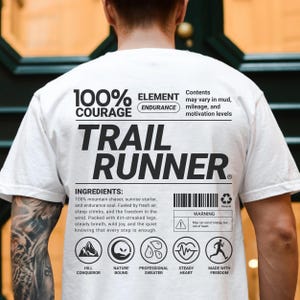 Op de afbeelding: Wit T-shirt met de tekst "TRAIL RUNNER" in grote zwarte letters. Het shirt heeft ook de tekst "100% COURAGE" en "ELEMENT ENDURANCE". Onder de tekst staan ingrediënten- en waarschuwingslabels met pictogrammen.