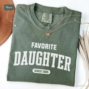 May include: A moss green t-shirt with the words "FAVORITE DAUGHTER" printed in white block letters, with "SINCE 1985" below. A silver necklace with a pendant rests on the shirt. The shirt is folded.