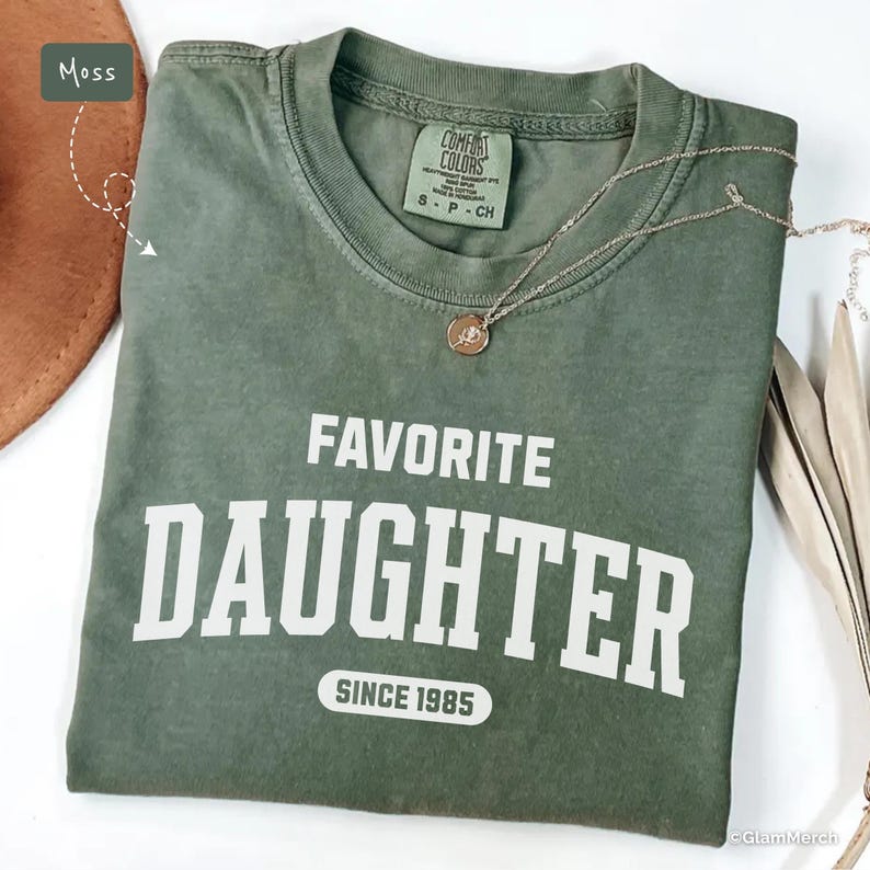 May include: A moss green t-shirt with the words "FAVORITE DAUGHTER" printed in white block letters, with "SINCE 1985" below. A silver necklace with a pendant rests on the shirt. The shirt is folded.