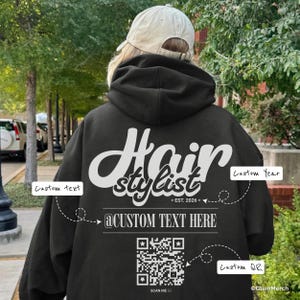 May include: A black hooded sweatshirt featuring white text on the back that reads Hair stylist, EST. 2026, and @CUSTOM TEXT HERE. Below the text is a square QR code with the words SCAN ME underneath. The hoodie is worn with a beige baseball cap.