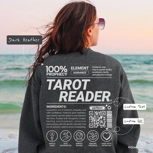 Könnte beinhalten: Dunkelgraues Sweatshirt mit dem weißen Text "TAROT READER". Auf der Rückseite des Sweatshirts befindet sich zusätzlicher Text, darunter "100% PROPHECY" und "ELEMENT GUIDANCE". Das Design enthält einen QR-Code und andere Grafiken.