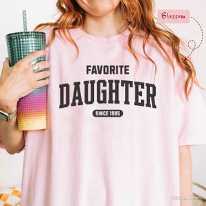 May include: A light pink t-shirt with the words "FAVORITE DAUGHTER SINCE 1985" printed in black. The shirt is being held by a person with a colorful tumbler. The tumbler has a green straw.