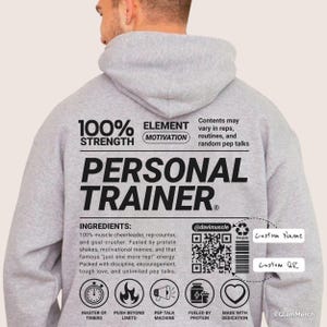 Peut inclure: Un sweat-shirt gris avec l'inscription "PERSONAL TRAINER" en grosses lettres audacieuses. Le dos du sweat-shirt présente une liste d'"INGREDIENTS" et des icônes. Le sweat-shirt porte également les mots "100% STRENGTH" et "ELEMENT MOTIVATION".