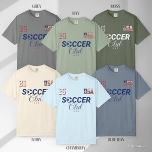 May include: A collection of six short-sleeve t-shirts in grey, bay, moss, ivory, chambray, and blue jean colors. Each shirt features the text '26', 'USA' in a flag pattern, and 'SOCCER Club' with a soccer ball graphic on the front chest area.