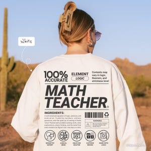 以下が含まれることがあります： 「MATH TEACHER」の文字が大きく黒字でプリントされた白いスウェットシャツ。背中には「100% ACCURATE ELEMENT LOGIC」の文字と材料リストも記載されています。クルーネックと長袖のデザインです。