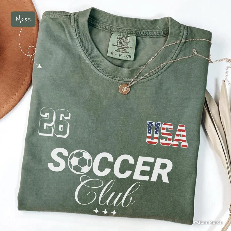 May include: A moss green short-sleeve t-shirt featuring the number 26, a soccer ball graphic, and the text "SOCCER Club" in white. The shirt also displays "USA" in a red, white, and blue flag pattern. A thin gold chain necklace rests on the collar.