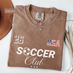 May include: An espresso-colored short-sleeve t-shirt with a Comfort Colors label. The front features the number 26, a USA flag graphic, and the text SOCCER Club with a soccer ball icon. A silver-toned necklace with a small pendant rests on the shirt.