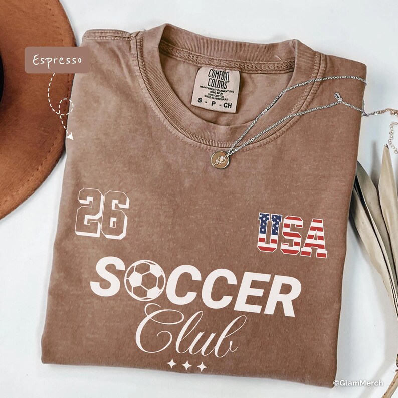 May include: An espresso-colored short-sleeve t-shirt with a Comfort Colors label. The front features the number 26, a USA flag graphic, and the text SOCCER Club with a soccer ball icon. A silver-toned necklace with a small pendant rests on the shirt.