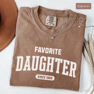 May include: Espresso-colored t-shirt with the words "FAVORITE DAUGHTER" printed in white, along with "SINCE 1985". A silver necklace with a pendant is draped across the shirt. The shirt is made of a soft material.