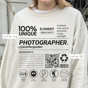 May include: Cream-colored sweatshirt with the word "PHOTOGRAPHER" and the text "100% UNIQUE" printed in black. The design includes icons and text about photography, a QR code, and the Instagram handle @jennifergarden.