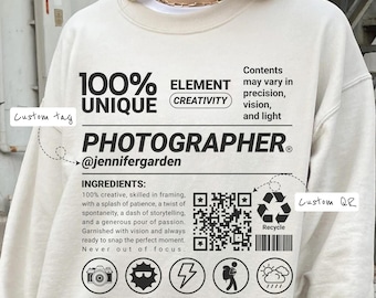 May include: Cream-colored sweatshirt with the word "PHOTOGRAPHER" and the text "100% UNIQUE" printed in black. The design includes icons and text about photography, a QR code, and the Instagram handle @jennifergarden.