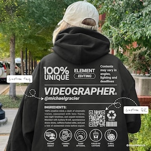Könnte beinhalten: Schwarzer Kapuzenpullover mit weißem Text: "100% UNIQUE VIDEOGRAPHER." Enthält eine Zutatenliste und Symbole. Der Hoodie hat ein individuelles Etikett und einen QR-Code. Der Text lautet auch "Editing Addict", "Camera Nerd", "Sleep Optional", "Fueled by Caffeine" und "B-Roll Junkie."