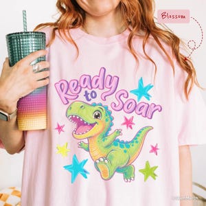 以下が含まれることがあります： 紫色の文字でReady to Soarと書かれ、カラフルな星に囲まれた緑色の恐竜のグラフィックが描かれたライトピンクのTシャツ。Tシャツには、ティールからオレンジへと変化するグラデーションカラーのテクスチャ付きタンブラーが添えられています。