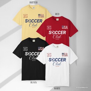 May include: Four short-sleeve t-shirts in butter yellow, red, black, and white. Each shirt features the text 26, USA, and SOCCER Club with a soccer ball graphic. The USA text is styled with a stars and stripes pattern, and three small red stars are centered below the word Club.