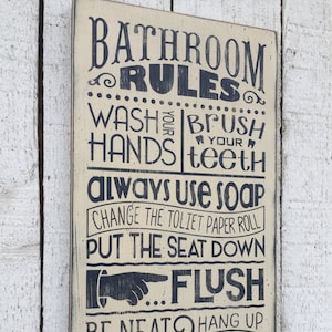 May include: A rustic wooden sign with a cream background and black text. The sign reads "Bathroom Rules" and lists a set of rules for bathroom etiquette, including "Wash your hands", "Brush your teeth", "Always use soap", "Change the toilet paper roll", "Put the seat down", "Flush", "Be neat & tidy", "Hang up your towel", and "Turn off the light".